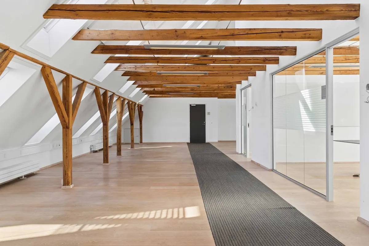 Lyst kontorlokal med lyse loftsbjælke og glasvinduer uden for. Loftsbjælken er lavet af træ og gulvet er lavet af træ. Lokalet er møbleret med skrivebord og stol.