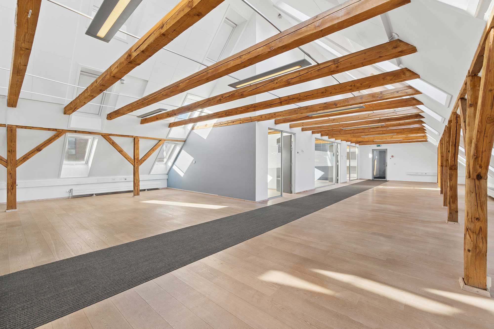 Kontorloft med lyse træbjælker, hvide vægge, gulve og lofter samt gulvbelægning og døre i baggrunden