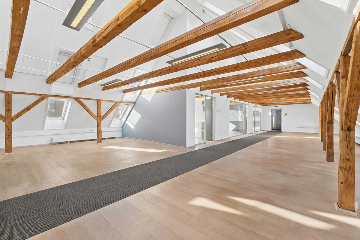 Kontorloft med lyse træbjælker, hvide vægge, gulve og lofter samt gulvbelægning og døre i baggrunden