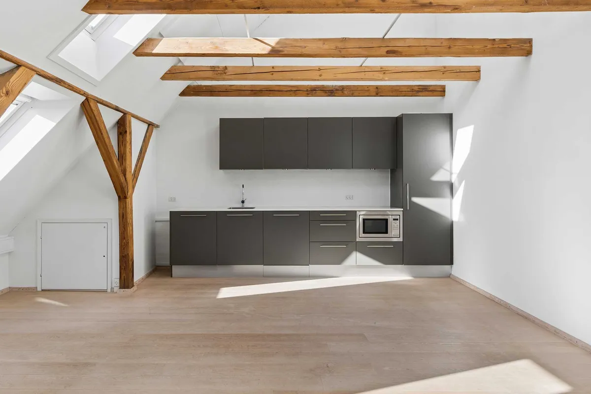 Et moderne loftsværelse med lyse gulv og højt loft med træbjælker, en komplet køkkenafdeling med grå skabe og en mikroovn