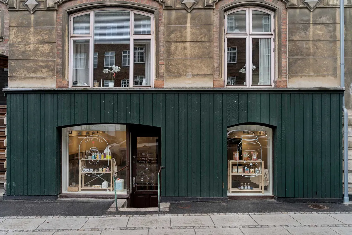 Store vinduer med hvide gardiner og grønne facader. Butik med ordet Refillery i vinduet og åbningstider markeret. Planter og mælkeglas i vinduet. Butikken ligger på en gågade.