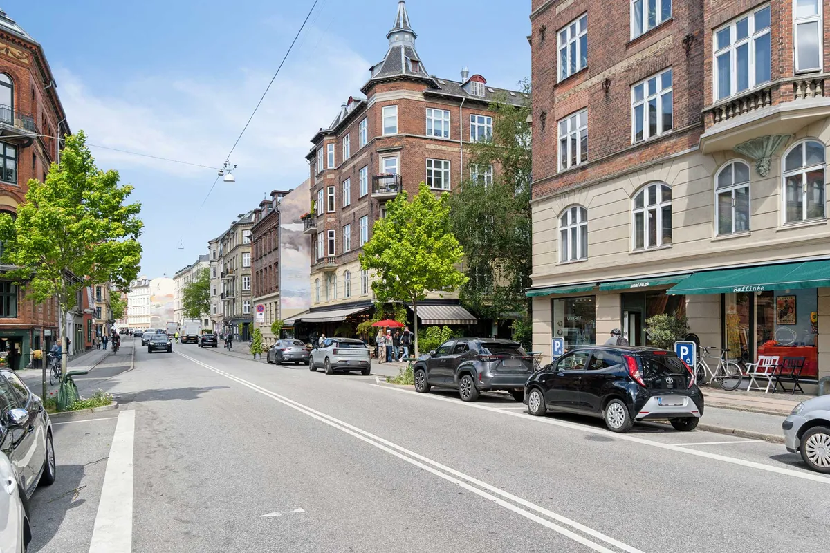 Biler parkeret langs gågaden med grønne træer og forretninger, der vender ud mod vejen med solrig himmel i baggrunden