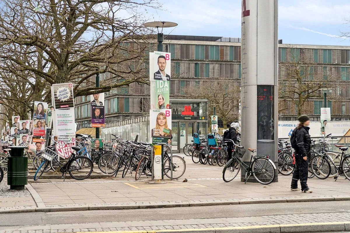 Foto af et cykelparkeringsområde med mange parkerede cykler og skilte med politikere på Frederiksberg. Foran står en person i sort og hvid tøj.