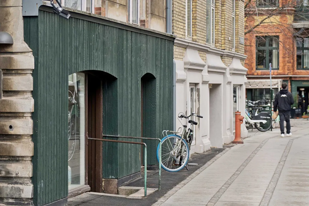 Mennesker går på gågaden foran en grøn facade med et enkelt vindue og sammen med en cykel og en hydrant