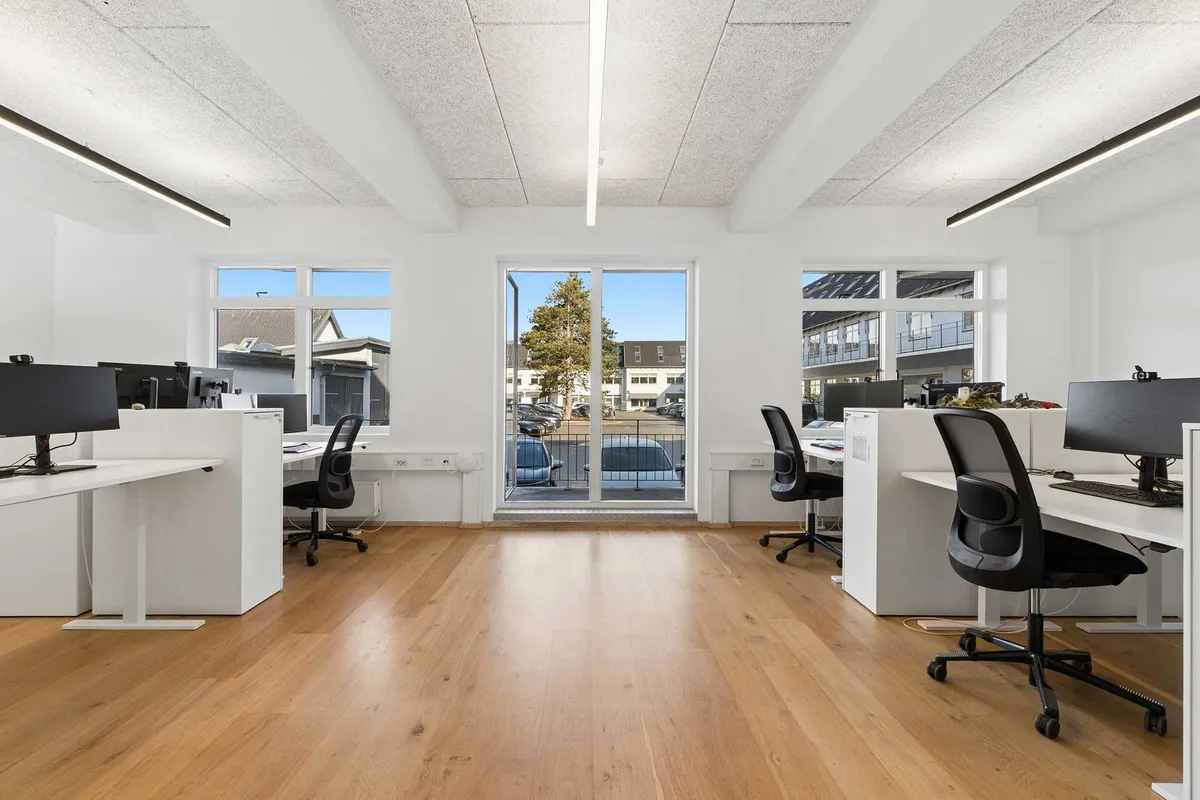 Kontorlejemål med højloftede rum og moderne indretning. Værelser med lyse gulve og hvide vægge. Kontorbord og stole i samme farve. Vindue med udsigt til parkeringsplads.