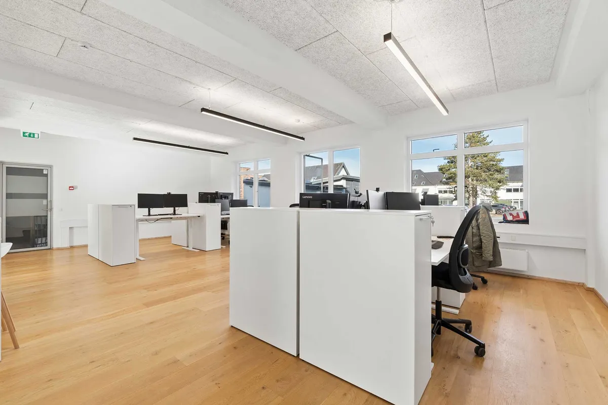 Kontorrum med højt til loftet og industrielle undertoner, lyse loftslamper, hvide vægge, trægulve, skriveborde, skærme, kontormøbler og ruder mod gårdhave med bygninger