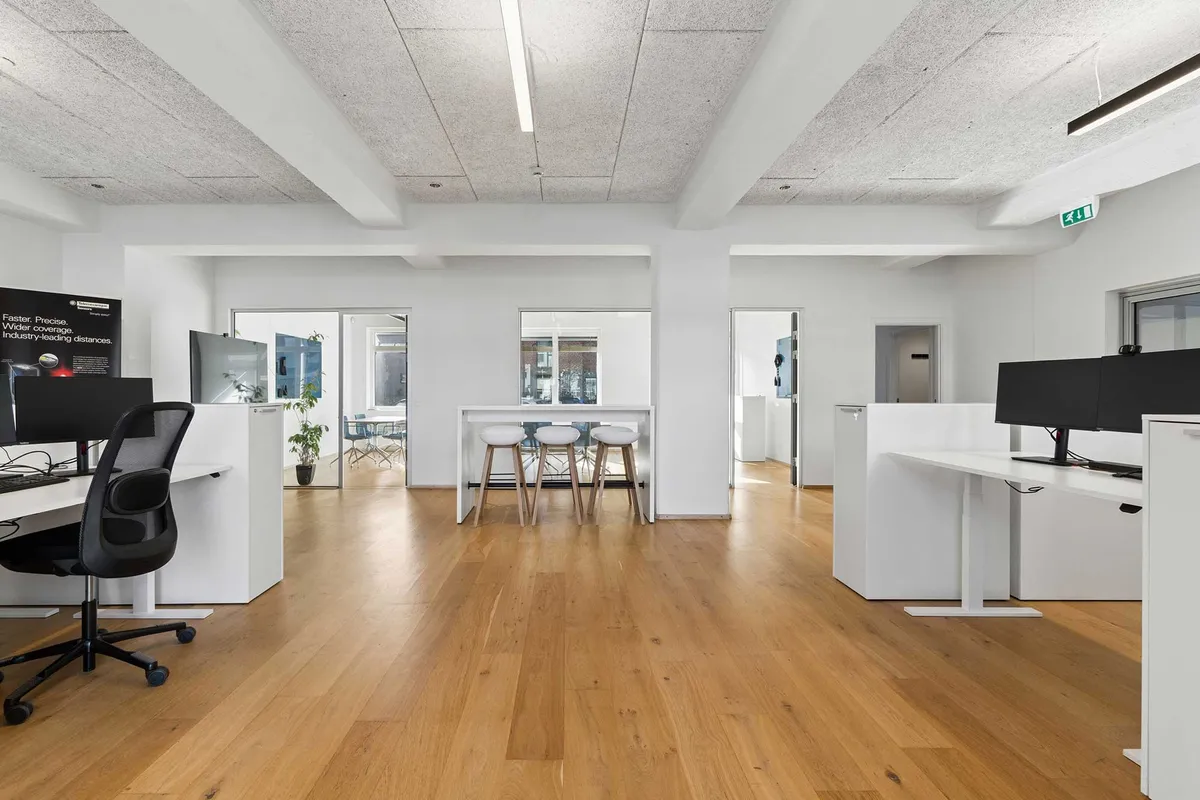 Stilfuldt kontor med højt til loftet og industrielementer. Hvidt møbeldesign, lyse hårde trægulve og moderne computerborde. To stueborde med fire høje stole og plante i baggrunden.