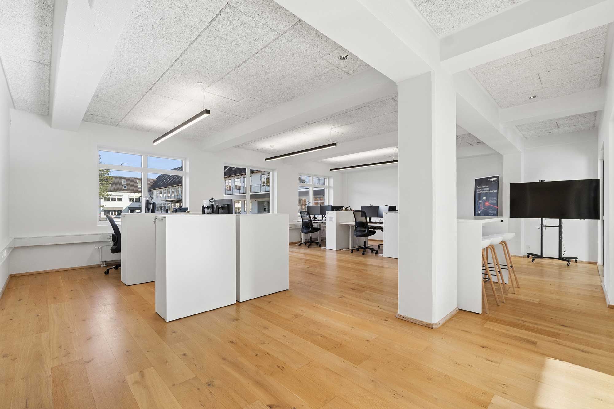 Kontor med højloft, lyse gulve og lyse skriveborde samt stolpe og monitorer.