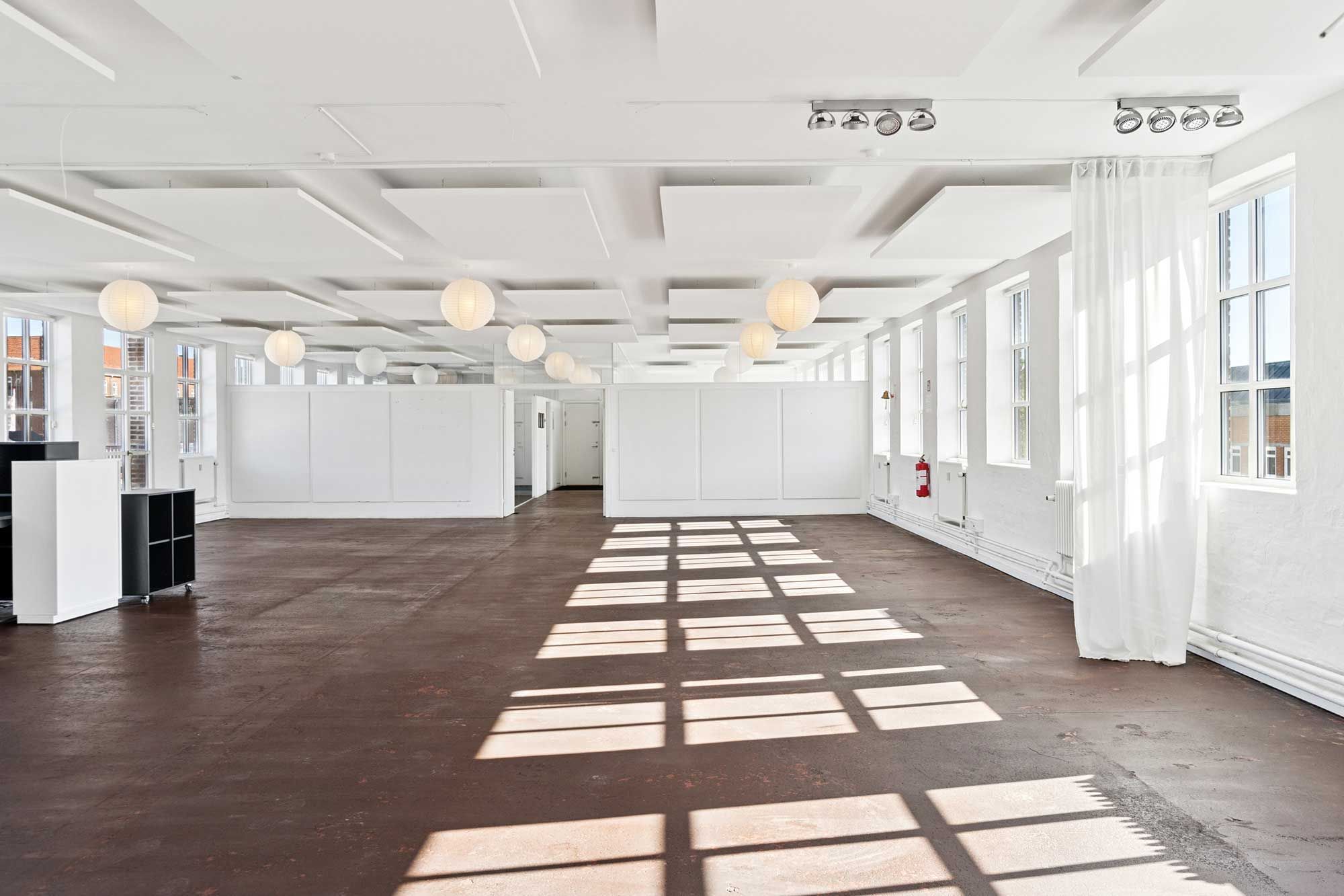 Råt kontorlejemål med store vinduer, hvide vægge og loftsbrædder, massivt lysindfald fra vinduer og lanterniner
