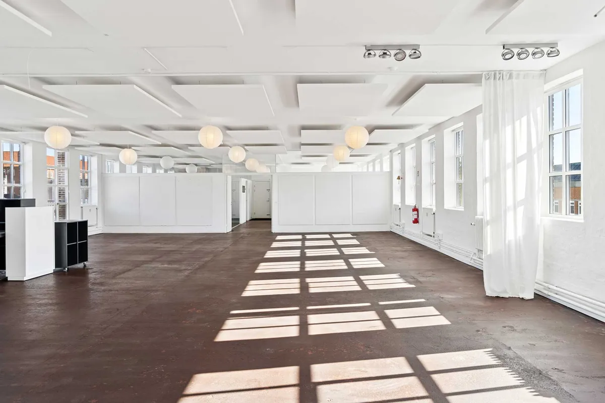 Råt kontorlejemål med store vinduer, hvide vægge og loftsbrædder, massivt lysindfald fra vinduer og lanterniner