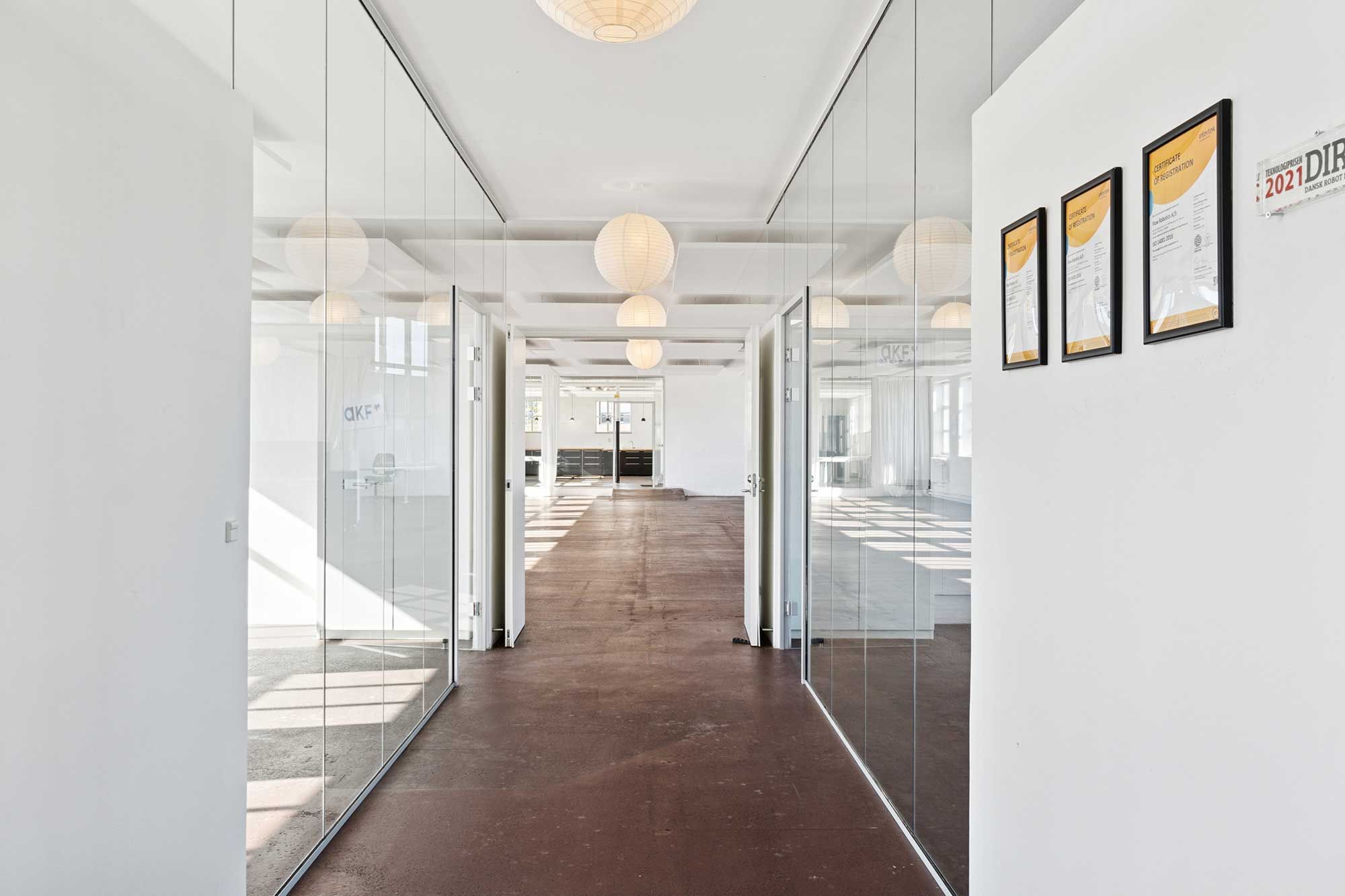 Rå kontorlokaler med massivt lysindfald, glaspartier og lyse loftslamper. Væggene er hvide, og der er tre certifikater hængende på en væg.