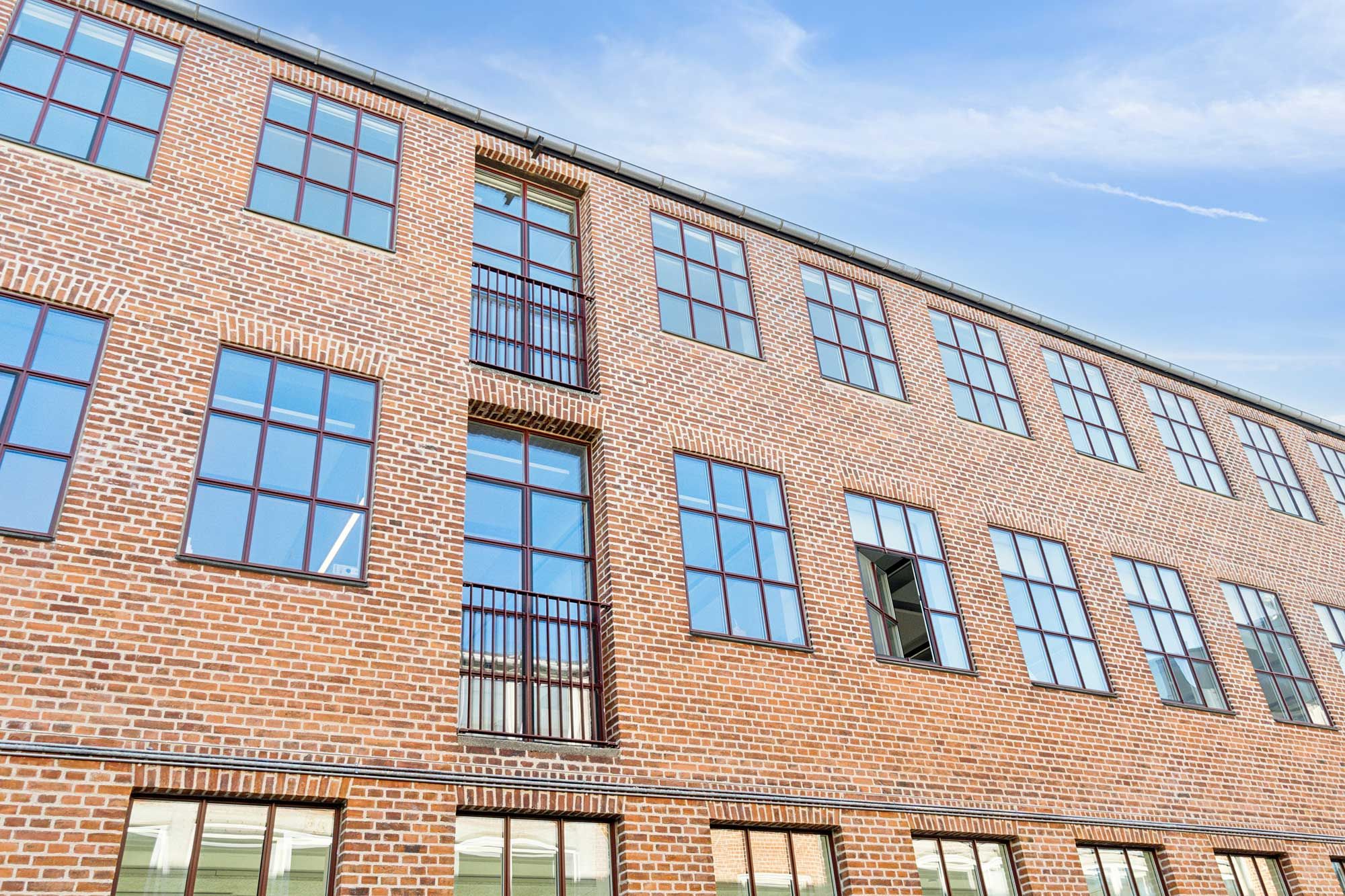 Råt kontorlejemål med store vinduer og røde mursten. Industrigulv og massivt lysindfald trænger ind i de store lokaler. Mange vinduerne er åbne.