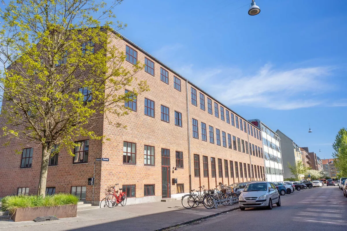 Kontorby i rå teglsten med masser af vinduer og flere cykler parkeret udenfor. Bygningen har lyserøde vægge og flere træer med lyse grønne blade.