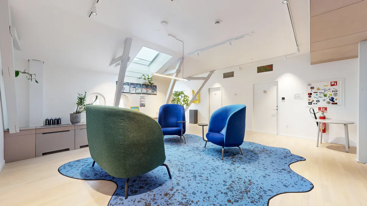 Kontorlokaler med lyse møbler og loftsrum med loftsvinduer. Væggene er hvide og der er store vinduer. Loftet er højt og lysekroner er installeret.