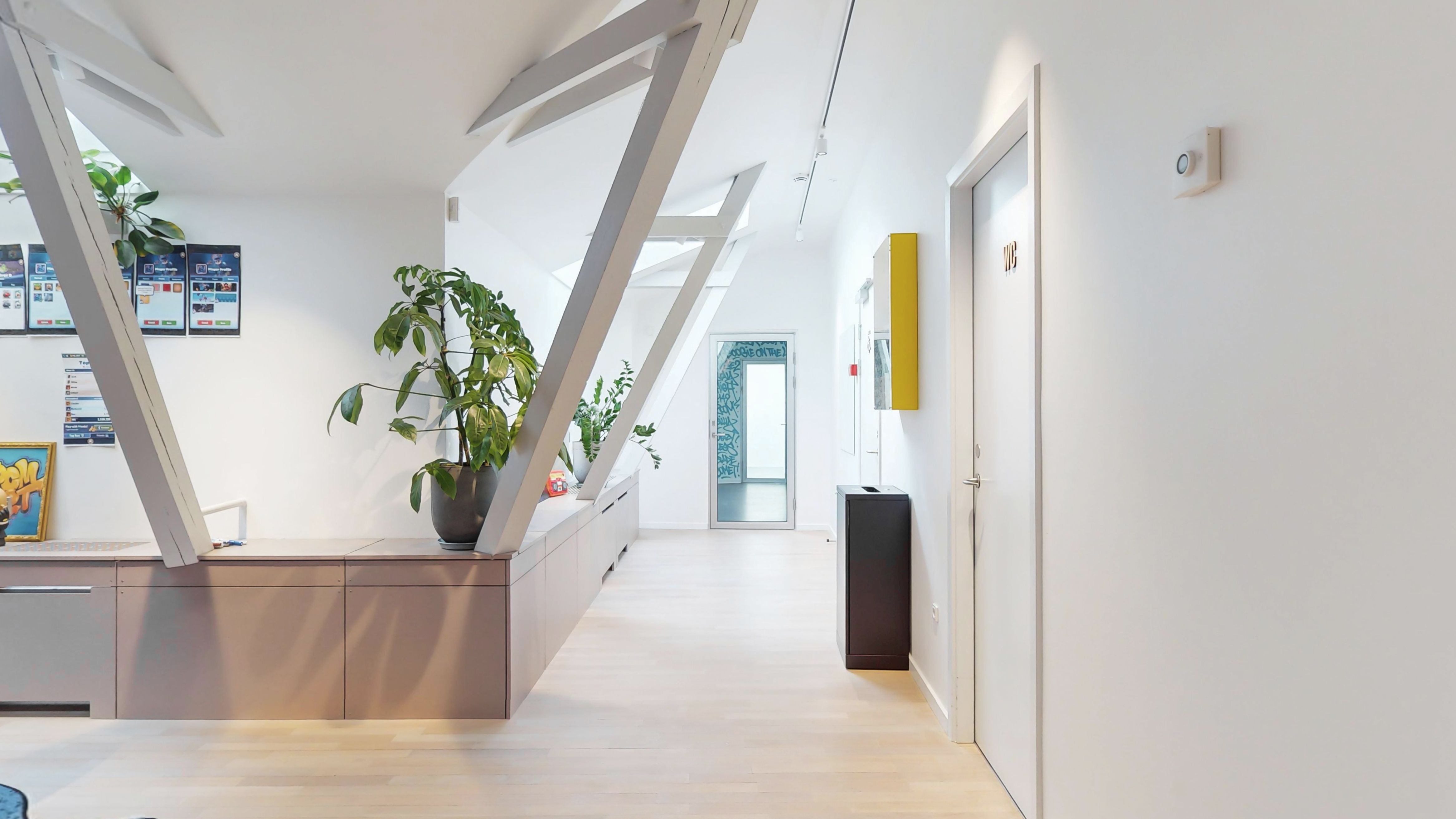 Kontorrum i baghuset med hvidt loft, loftsbjælker og lysekroner. Komplet med lysegul væg og en stor plante i lysebrunt potte. Rummet er møbleret med bord og stole