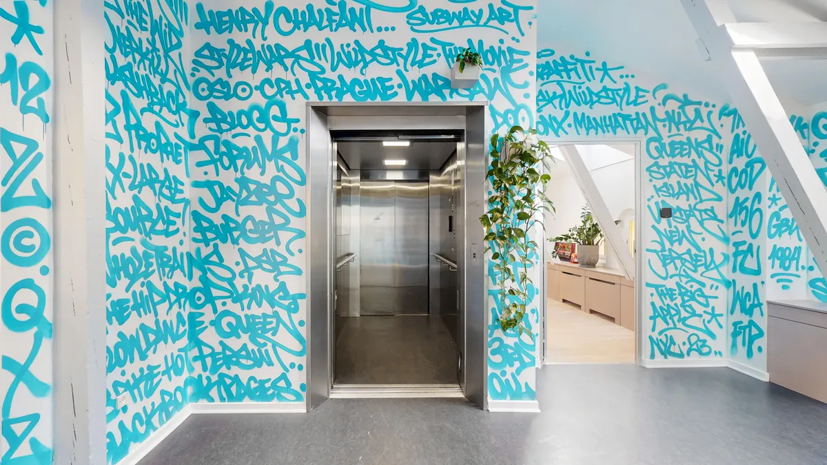 Kontorfremlejemål med graffiti på væggene og åbent elevator. Der er et nyt skrivebord med plante på. Loftet er højt og der er et nyt køkken med lyse skuffer