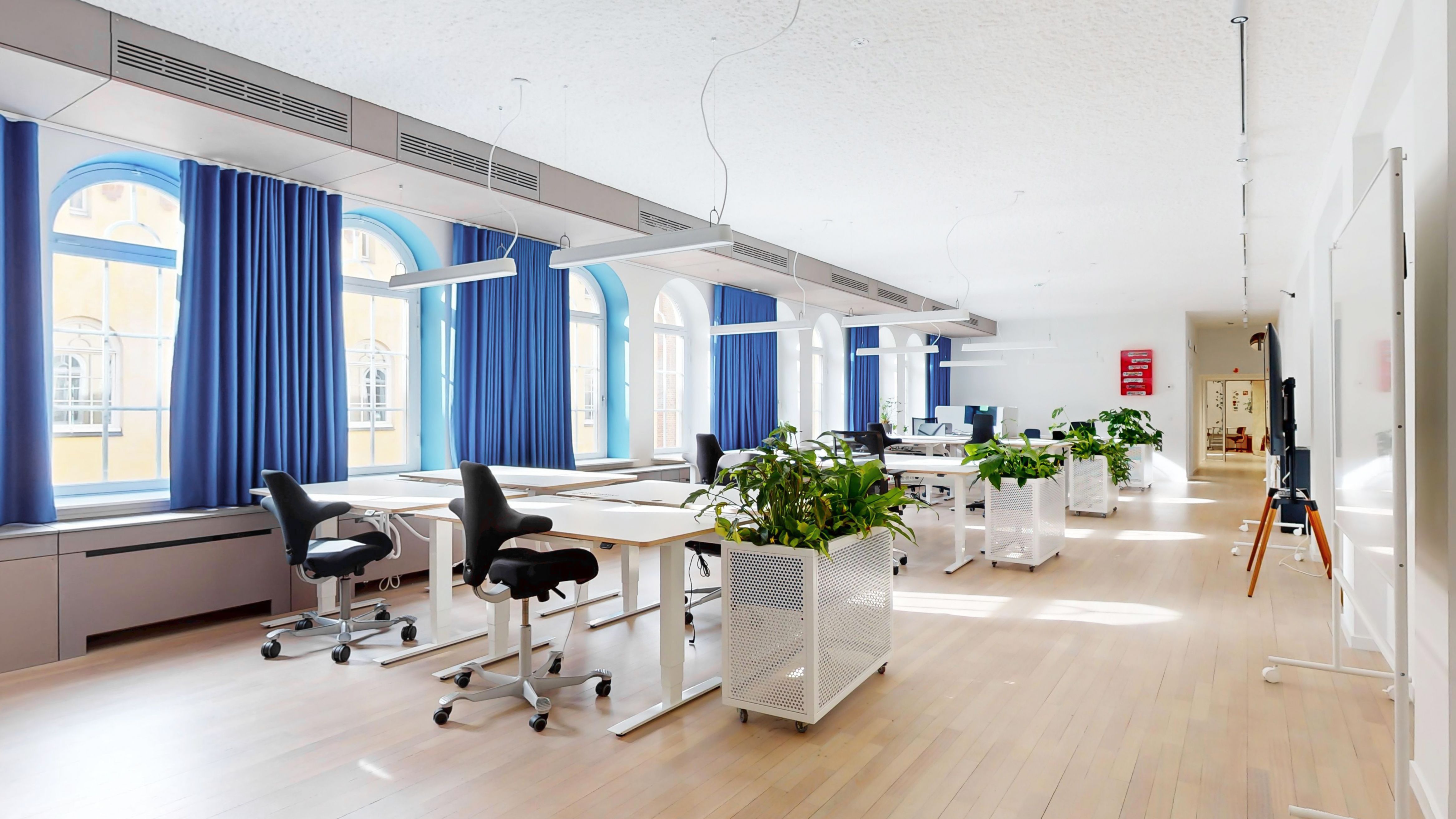 Kontorlokation med hvide gulve, lyse vinduer og blå gardiner. Møbler inkluderer hvide skriveborde, ergonomiske sædestole og hvide plantepoteter.