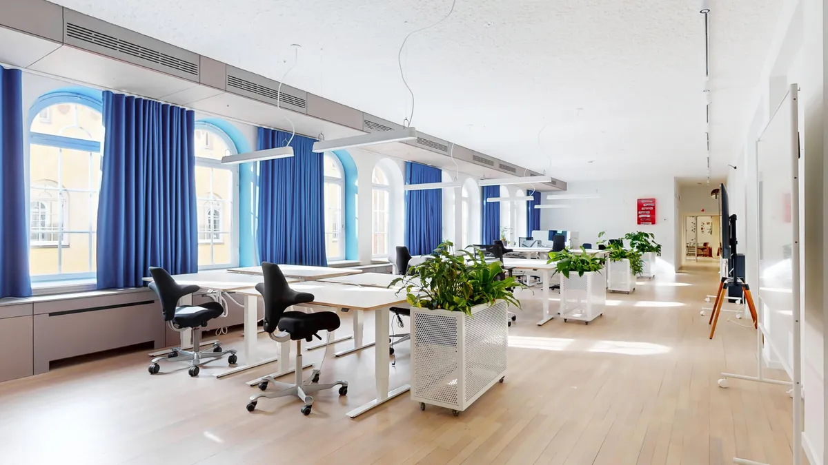 Kontorlokation med hvide gulve, lyse vinduer og blå gardiner. Møbler inkluderer hvide skriveborde, ergonomiske sædestole og hvide plantepoteter.