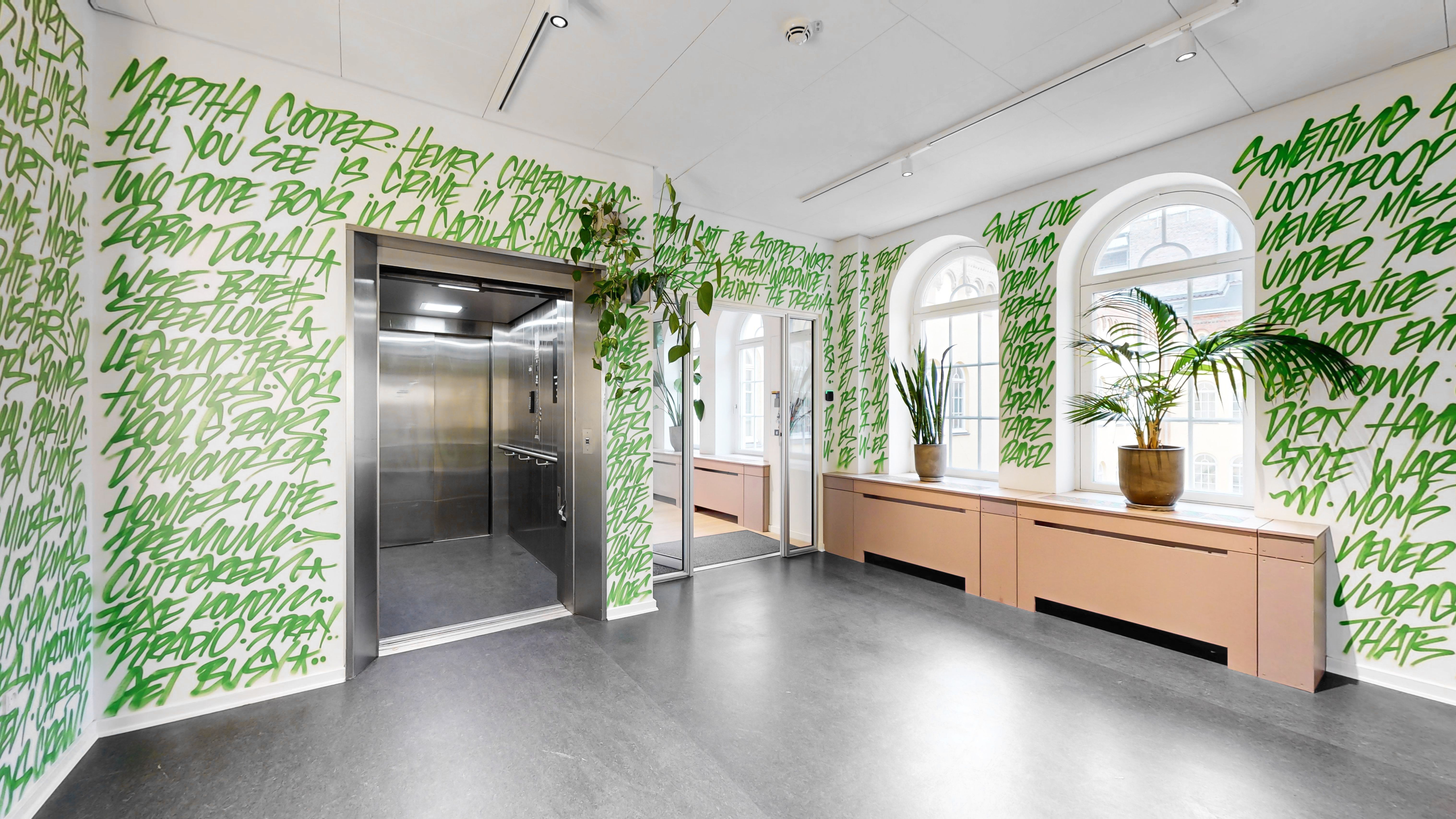 Kontorrum med elevator, lyse og lysegulve, og lysegrå og grønne vægmalerier. Vinduerne med bueformet ramme, og to store planter i potter på vindueskarmerne