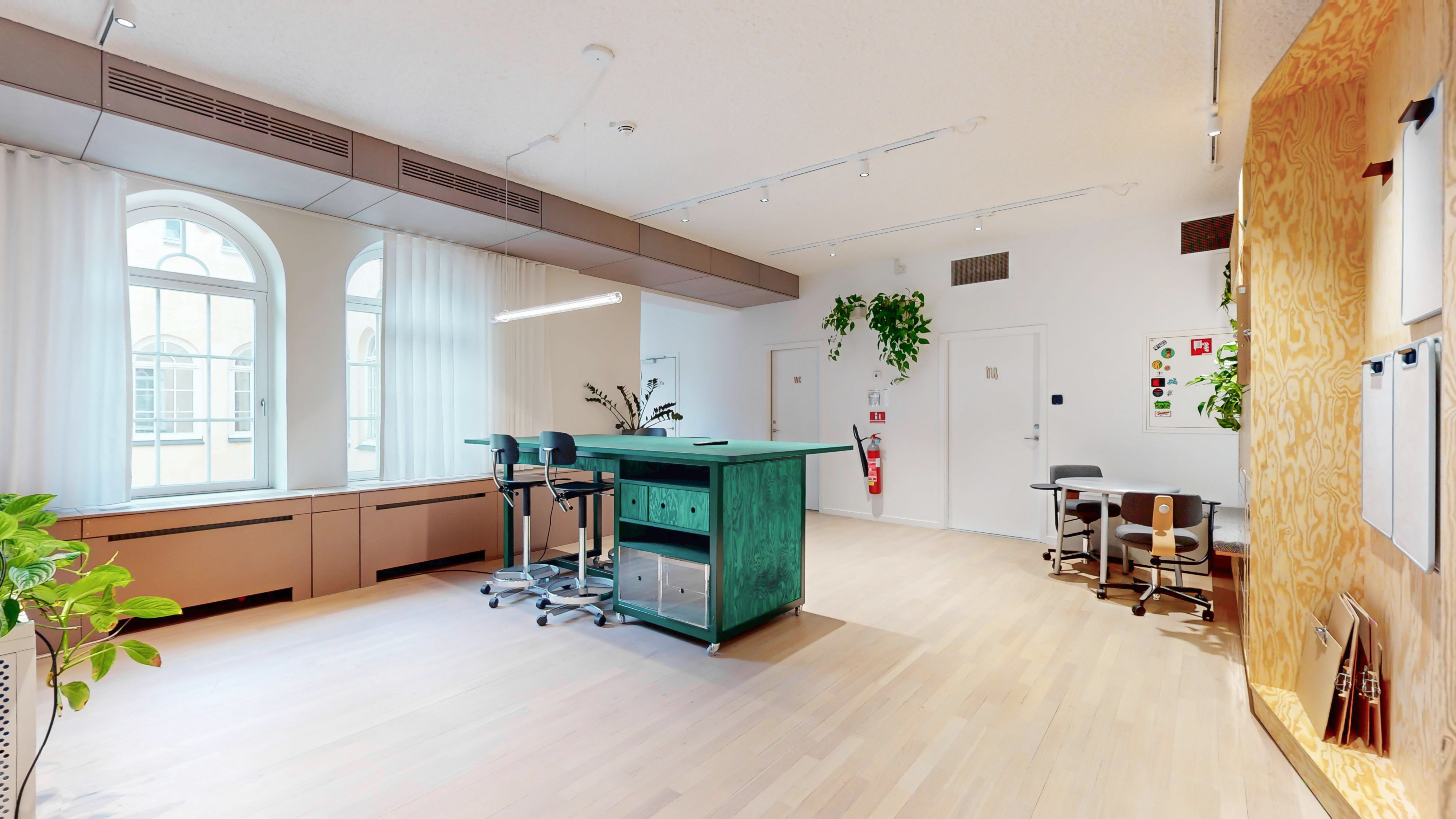 Kontorrum med højt loft og lyse vægge. Et stort grønt bord og høje stole til møder. Planter og hængelampe forbedrer kontorlokalets atmosfære.