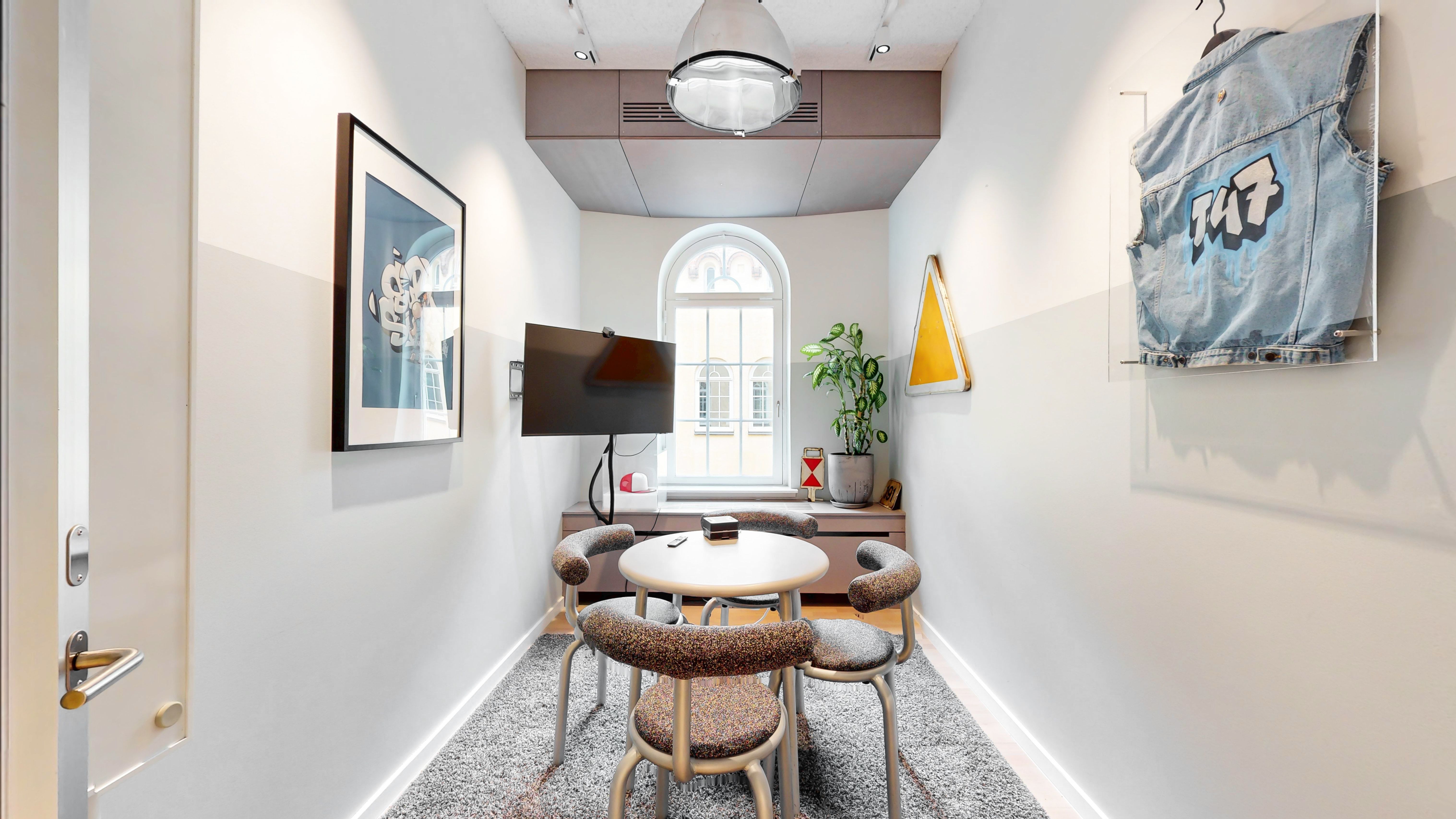 Kontorfremlejemål med runde spisebord, stolpe med jeansjakke og gult skilt, TV og plante i gulvet