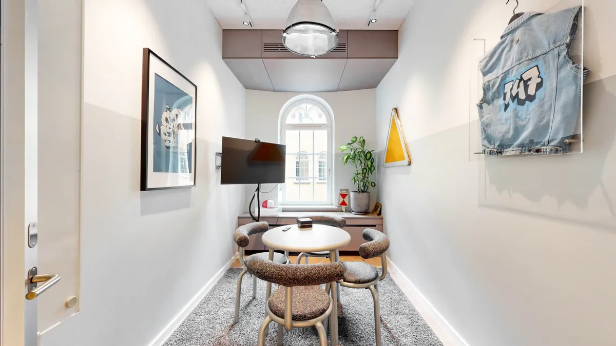 Kontorfremlejemål med runde spisebord, stolpe med jeansjakke og gult skilt, TV og plante i gulvet