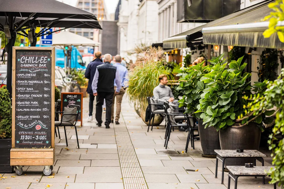Kontorlokaler med menubord og plantager på en gågade med mennesker og et hjørne af en bygning med mørk aula