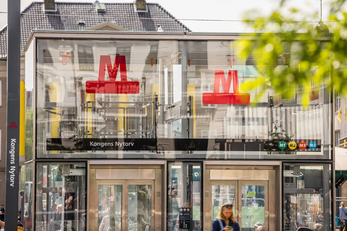 Modernt, glasklædt kontor med rødt M-logo og Kongens Nytorv-tekst i København. Bygningens refleksioner og grønne træer ser ud til at skabe en hyggelig arbejdsatmosfære.
