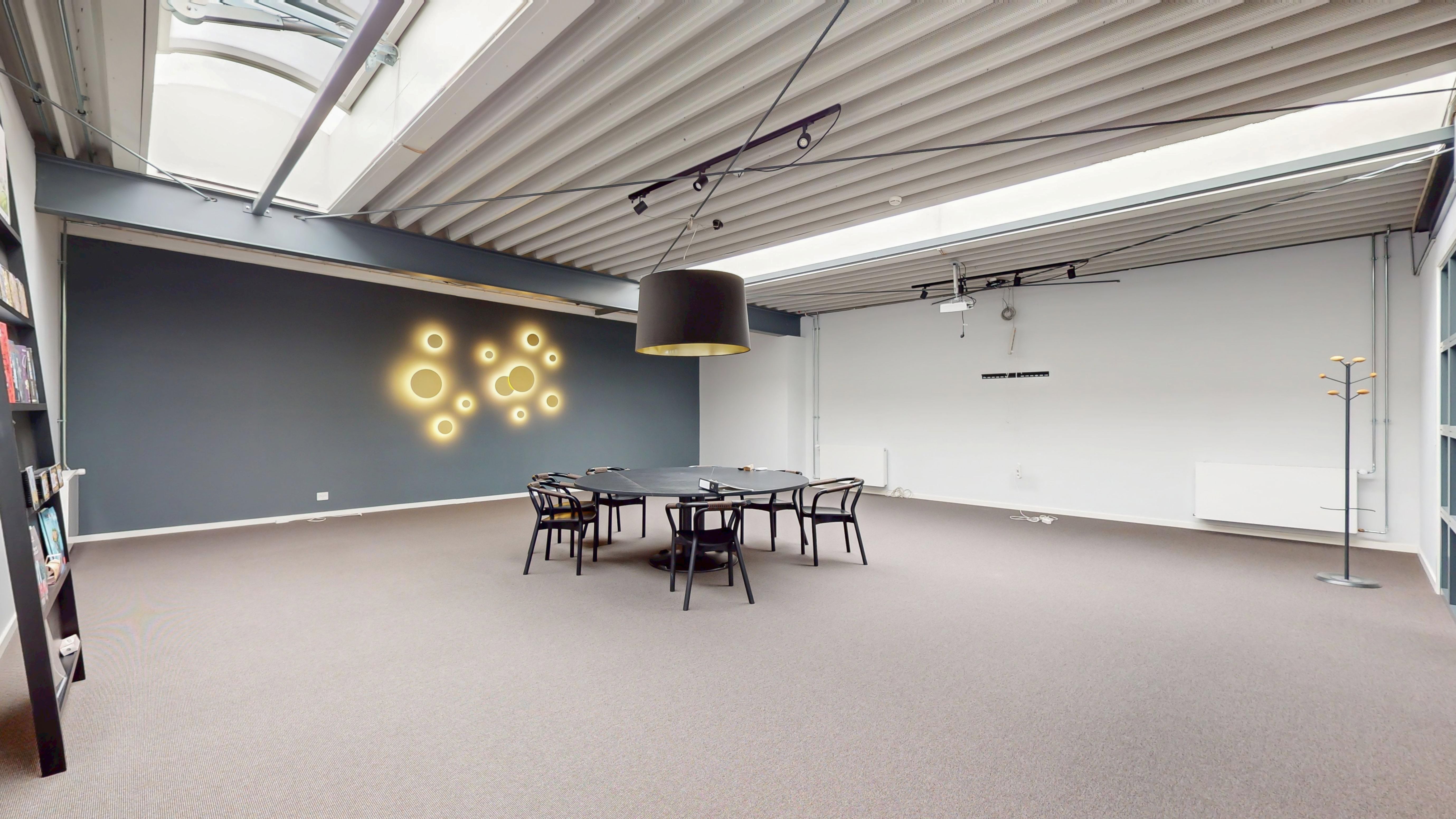 Et stort og fleksibelt kontorrum med hvide vægge, grå loft, runde gulvlampe og bord og stole samt en hylde med bøger