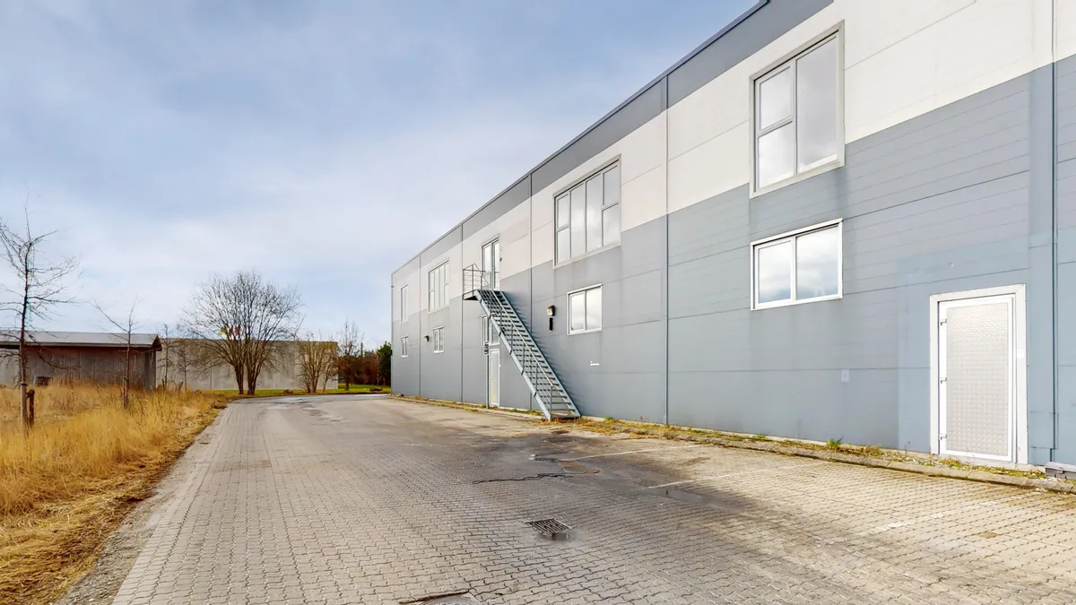 En stor bygning med vinduer og en trappe. Et område med snavset asfalt og søjler. En stribe træer uden løv og en anden bygning i baggrunden.