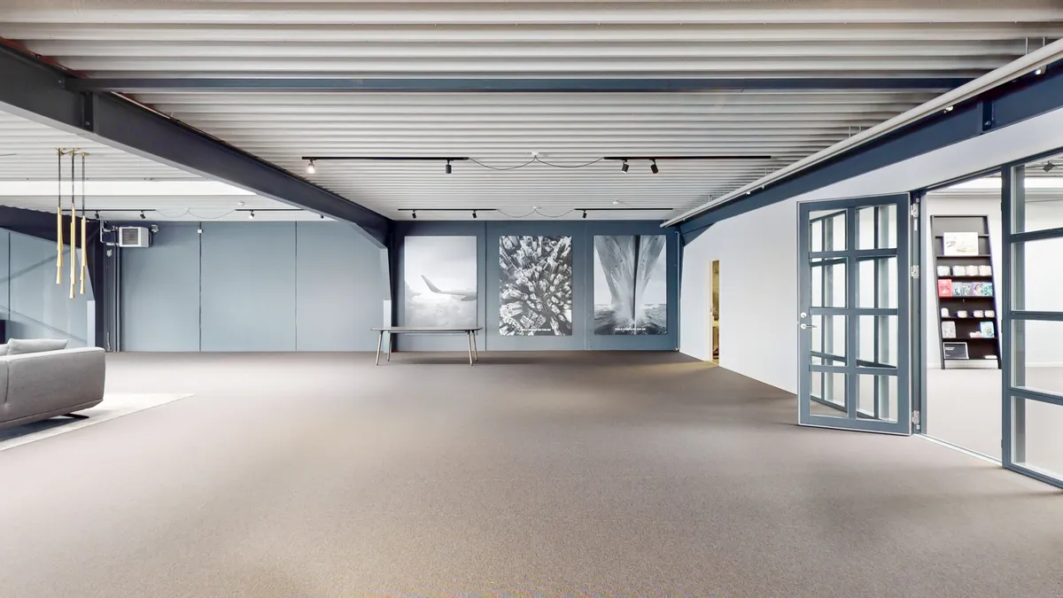 Det store kontor har åbent og fleksibelt gulvplan med lyse lofter og store vinduer. Væggene er lyse og minimalistiske og der er et stort, tomt rum med en stol og lyse gulvbelægning.