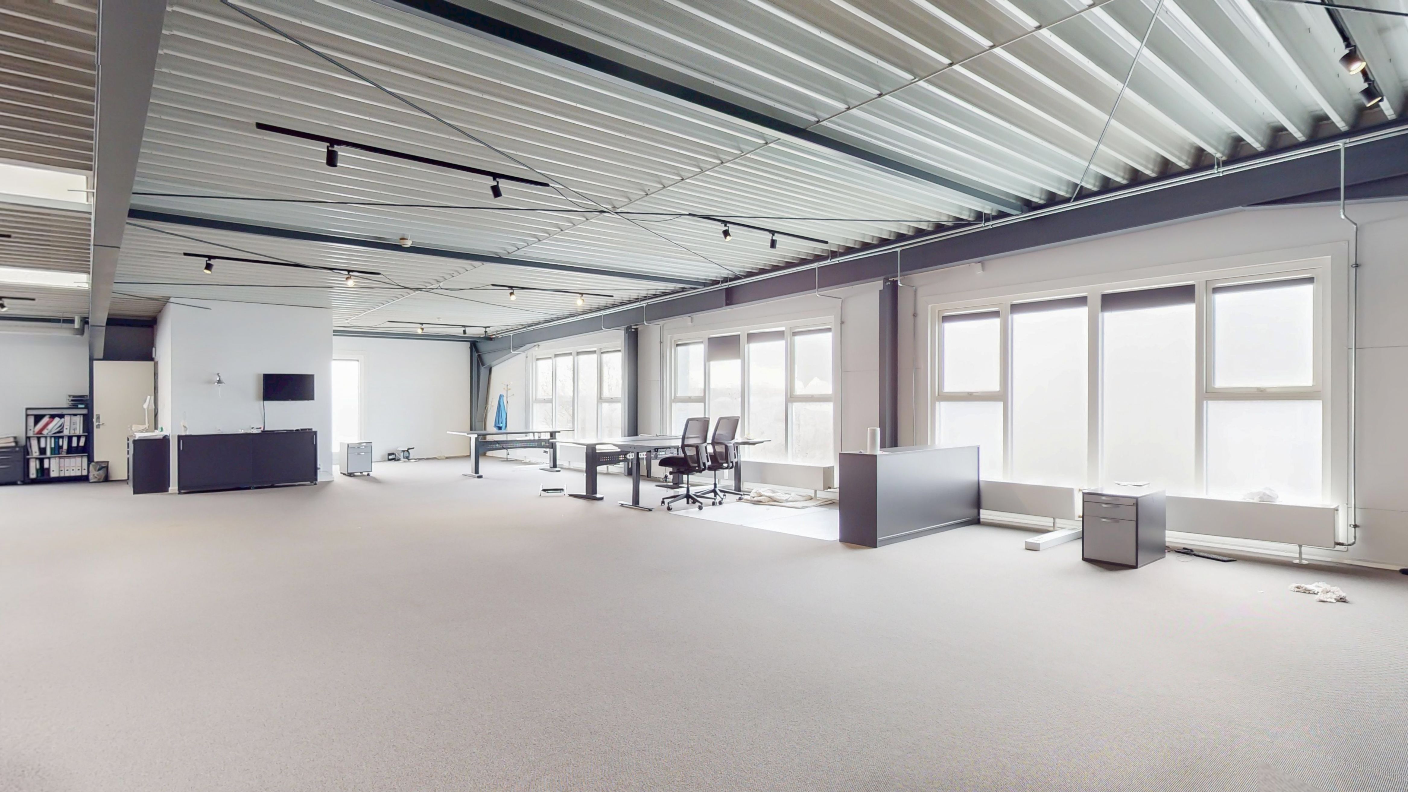 Stort kontorområde med lyse lofter og mange vinduer. Moderne møbler og teknologi er placeret i området. Gulvet er lyse og fladt.