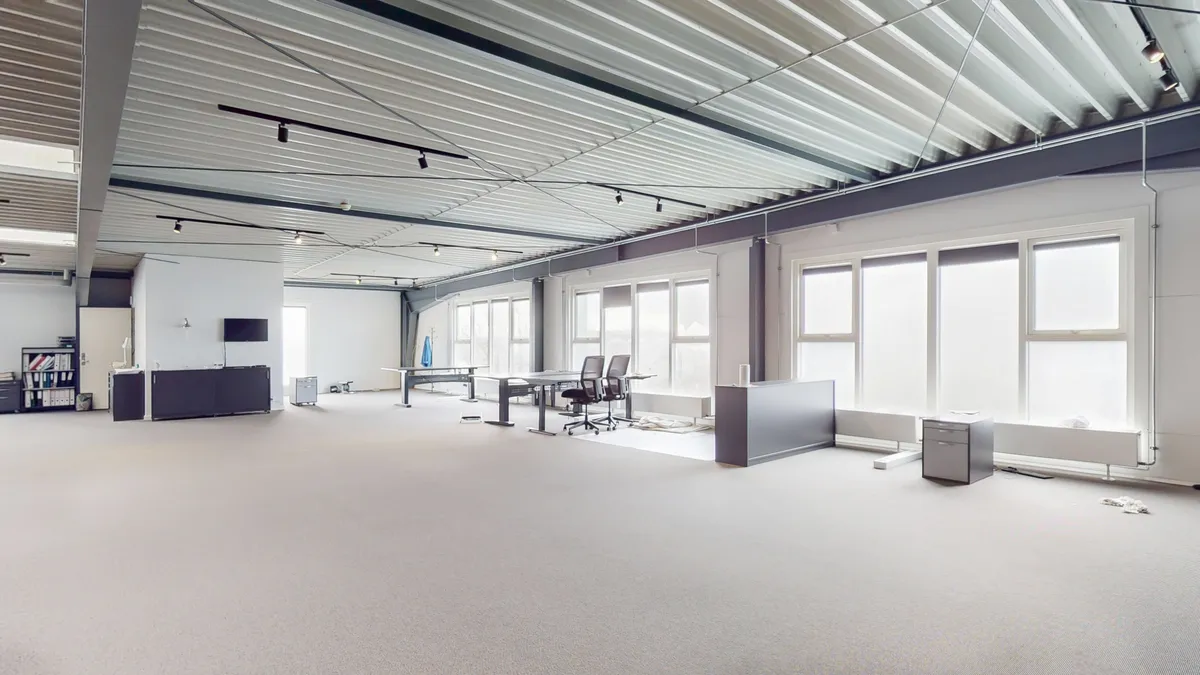 Stort kontorområde med lyse lofter og mange vinduer. Moderne møbler og teknologi er placeret i området. Gulvet er lyse og fladt.