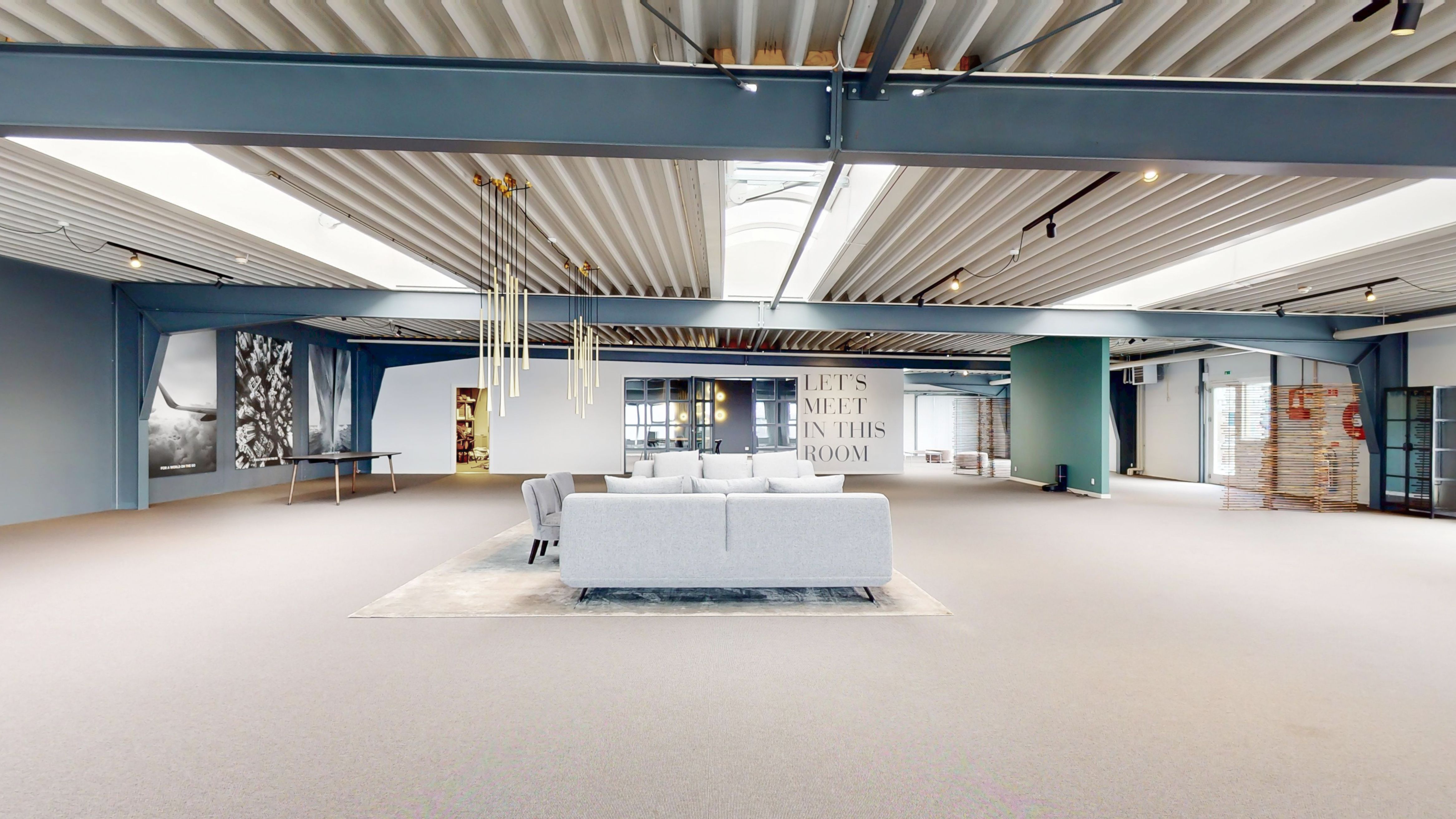 Fremlejemål på Stamholmen 140A med loftsvinduer og indvendig lysekroner. Minimalistisk interiør med møbelgruppe, tekstiler og gulvbelægning. Tekstskiltet "Let's Meet in This Room" står fremtrædende.