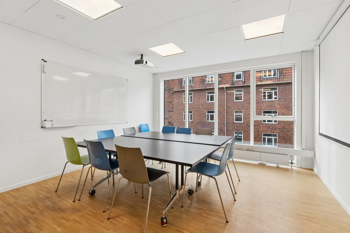 Kontorlokaler med mødesal og spisestue. Hvid væg med projektionsapparat og tavle. Farverige stole og spisestolsbord med hjul. Vindu med udsigt til røde bygninger.