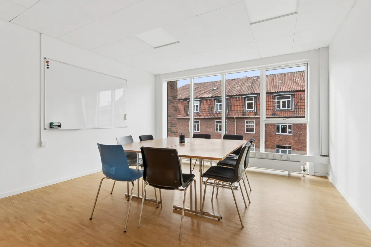 En mødelokale med lyse vægge, trægulv og store vinduer med udsigt til bygning. Væggen med en whiteboard og en række stole. Et skrivebord med en kuffert og en kop.