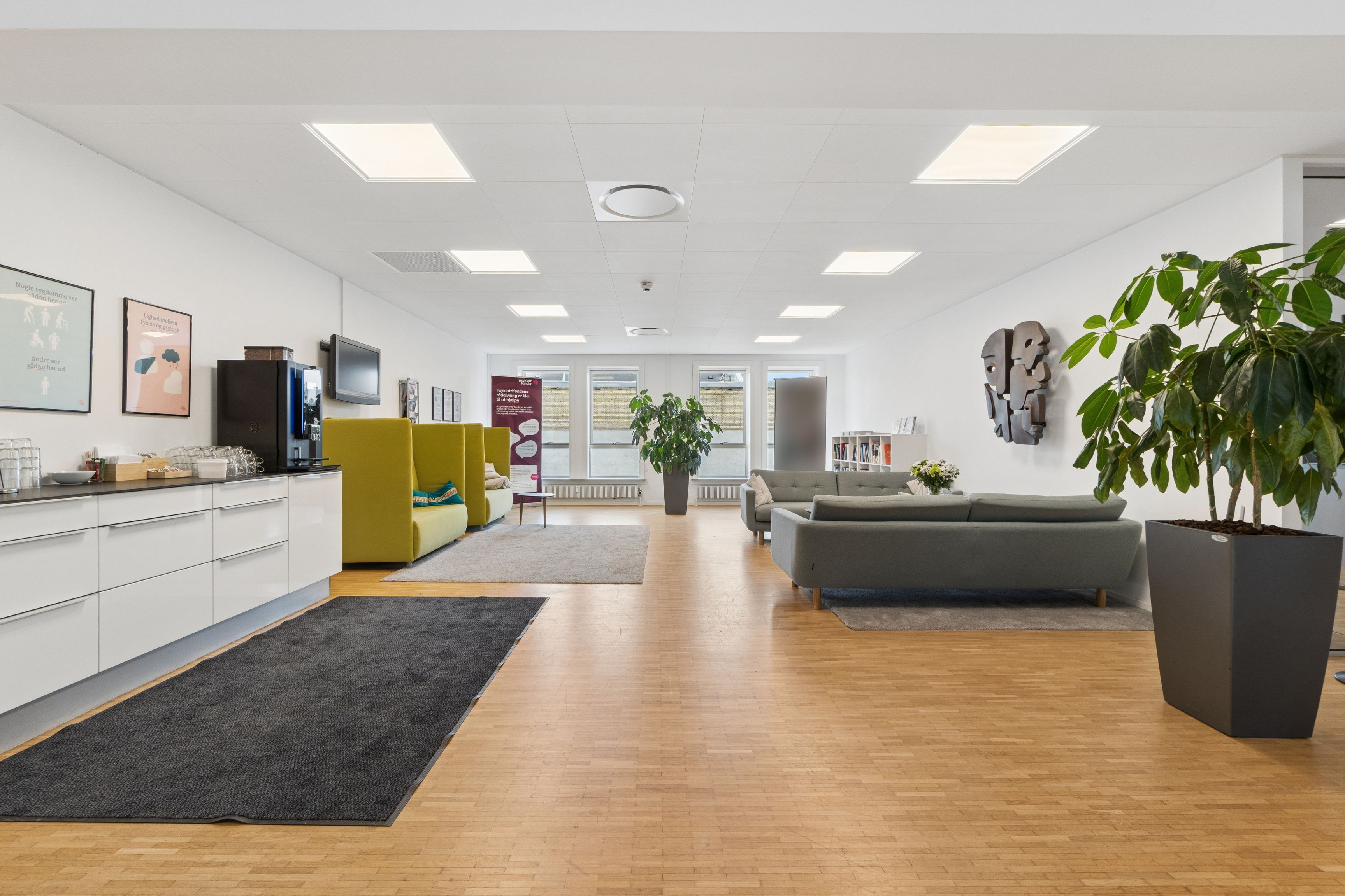 Nyistandsat kontorlejemål med lyse loftslofter og moderne møbler. Et moderne køkken med hvide skabsfronter og en grøn sofa i lysegråt og gult. En stor grøn plante og kunstværker på væggen.