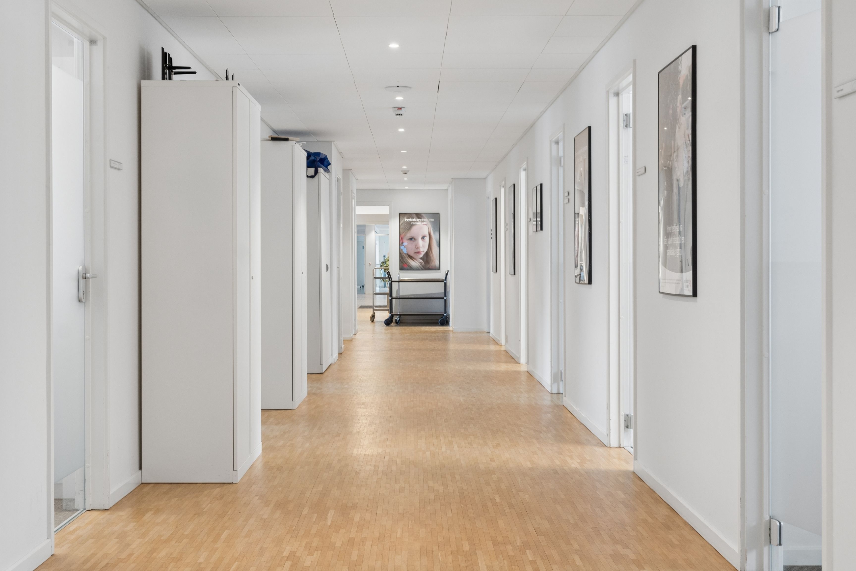 Lang korridor med hvide vægge og gulv af træ. Væggene er hvide og lukket med hvide døre. Der er flere vægmonterede billeder og reklamer.