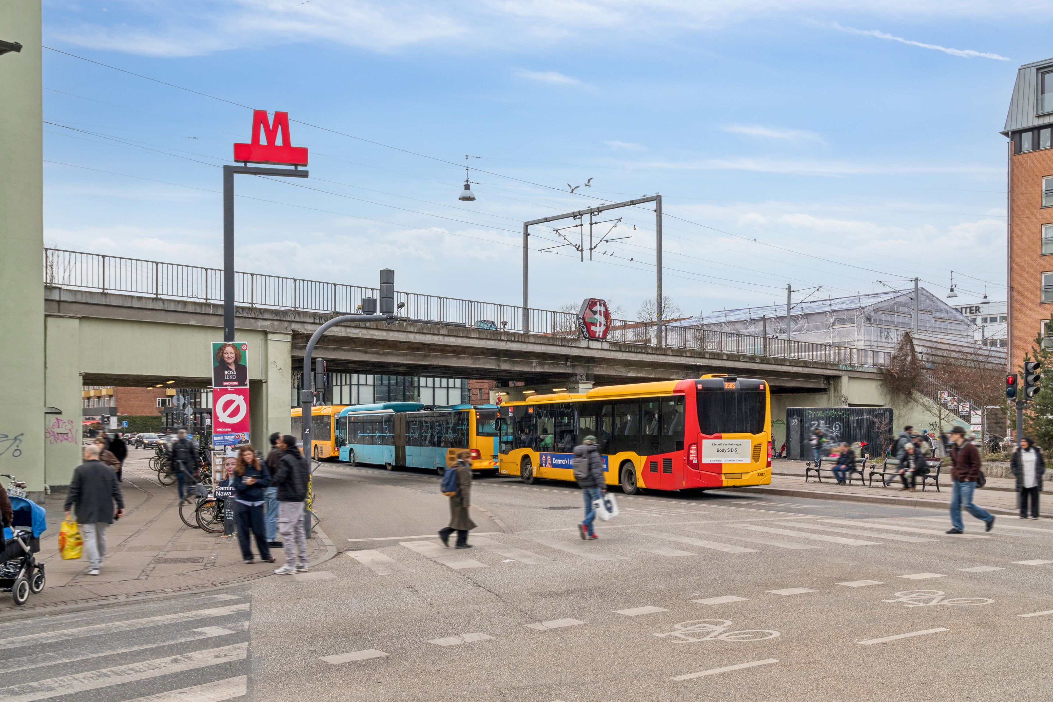 Flere mennesker krydser en gågade i København med flere busser i baggrunden og en metrostation i forgrunden med rødt M-skilt
