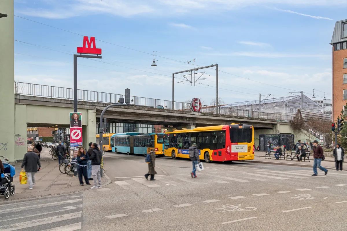 Flere mennesker krydser en gågade i København med flere busser i baggrunden og en metrostation i forgrunden med rødt M-skilt