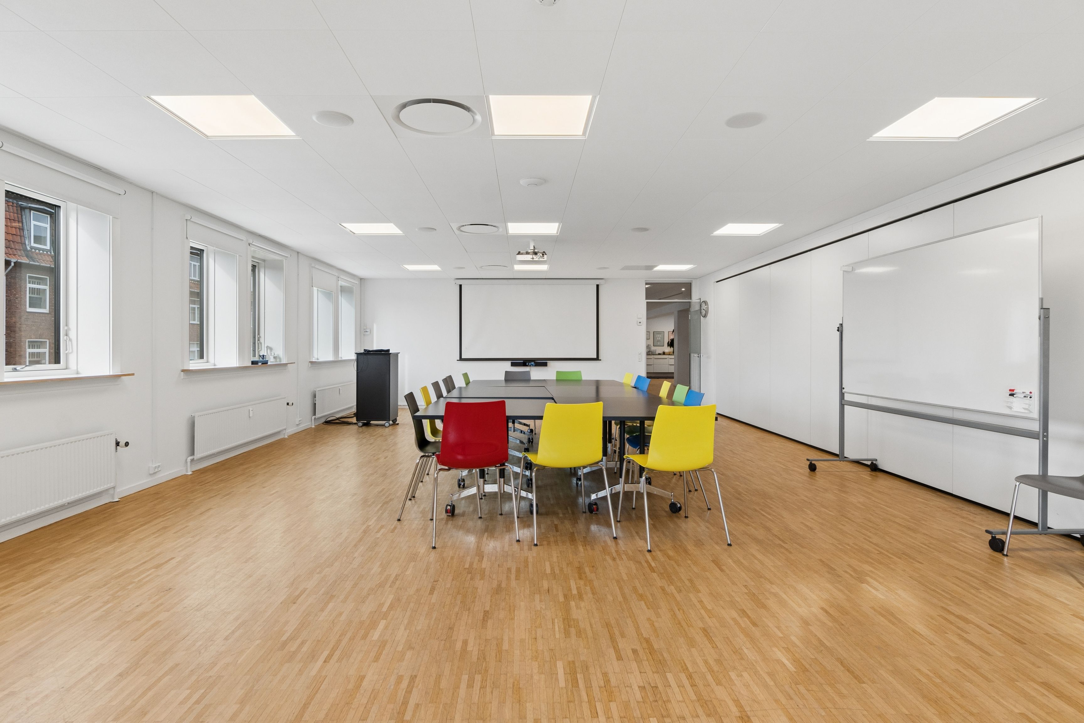 Kontorrum med trægulv og hvide vægge. Farverige stole og skrivebord samt hvidt skrivebord og projektor. Højt loft med flere indbyggede lys.