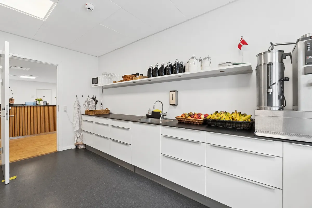 Hvidt køkken med højloft, moderne møbler og lysegulv, frugtkurv med æbler og bananer, to stålapparater og hvidt flag