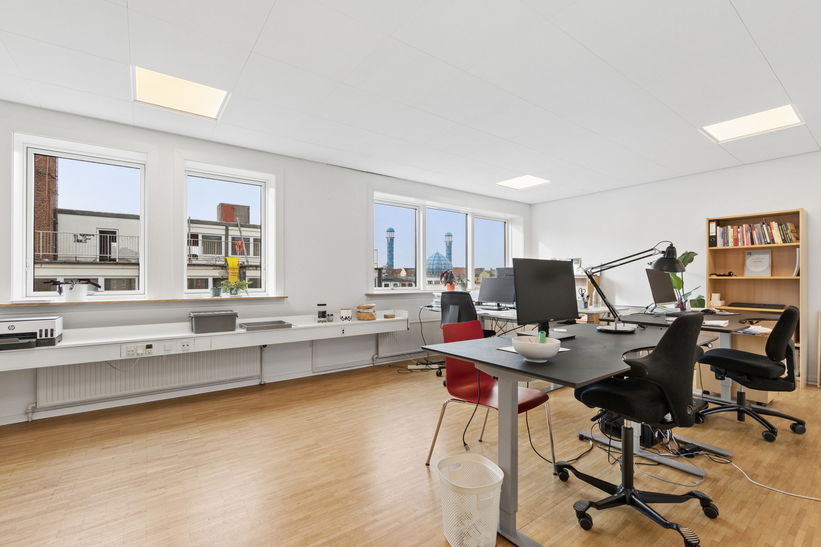 Lyst og indbydende kontor med moderne skrivebord, stol, computer og vinduer med udsigt til bygninger og tårne på afstand