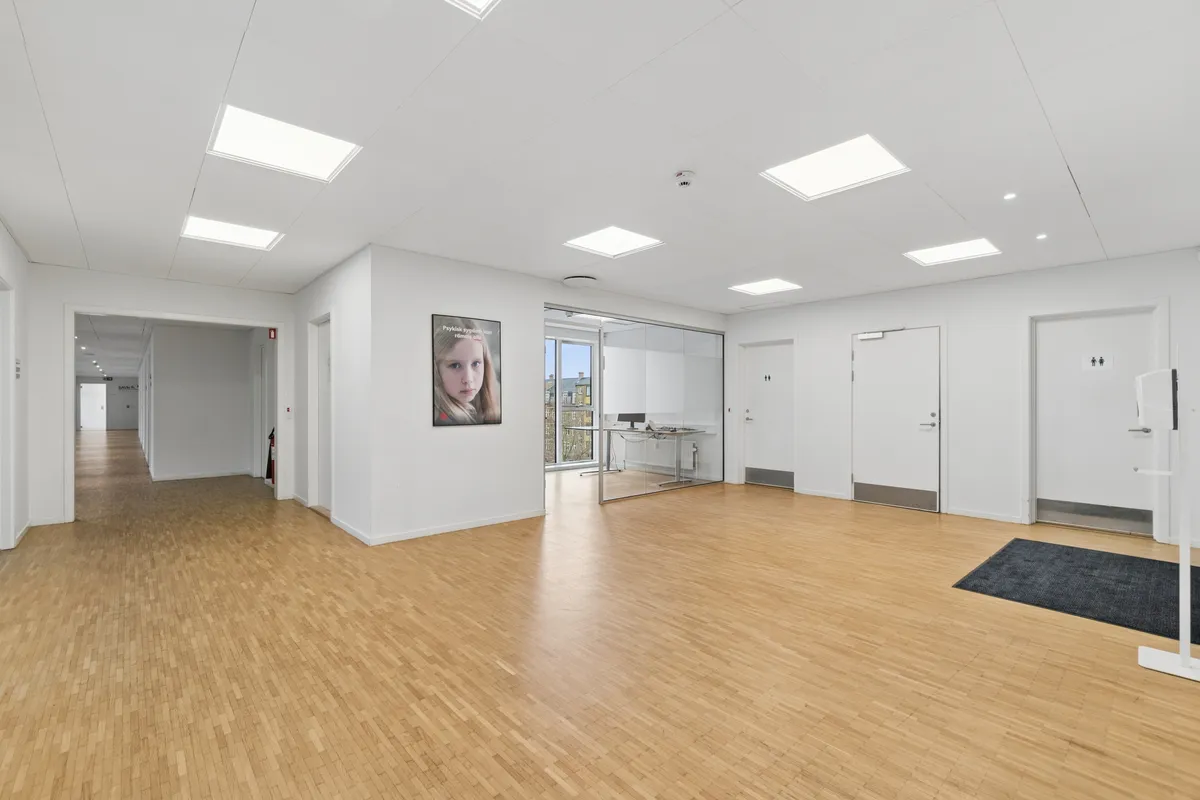 Et lyst og indbydende kontor med højt til loftet, store vinduer og masser af lys. Gulvet er lavet af træ og der er et lille værelse med en skrivebord og et billede på væggen