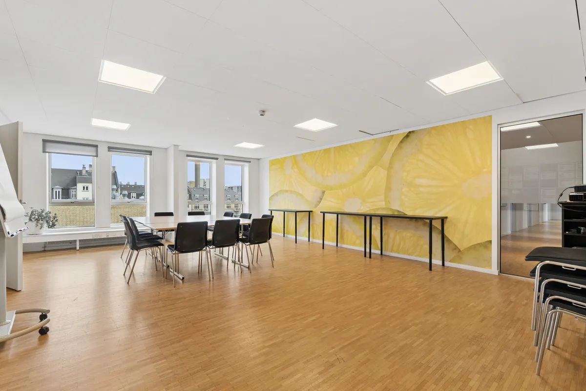 Lyst og indbydende konferencelokale med lyse vinduer og et stort gulv af træ. Bord og stole står klar til møder. Et lysegult vægmaleri med citronglimt og et lysegråt loft.