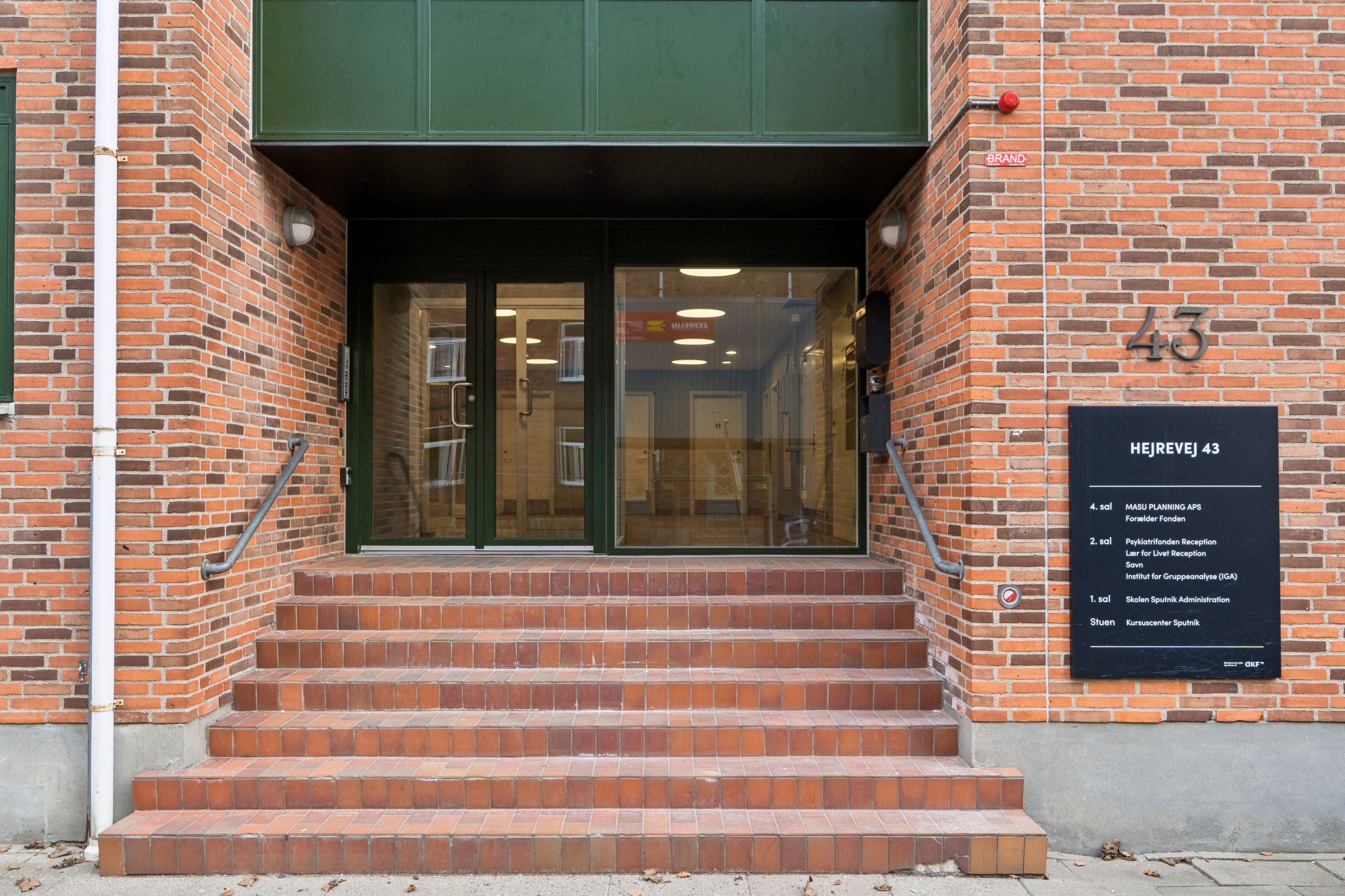 Kontorlejemål på Hejrevej 43 i København NV med lyse glasdøre og røde pudsede trin, som fører ind i bygningen. Der er et skilt med tekst, der angiver forskellige kontorer og faciliteter.