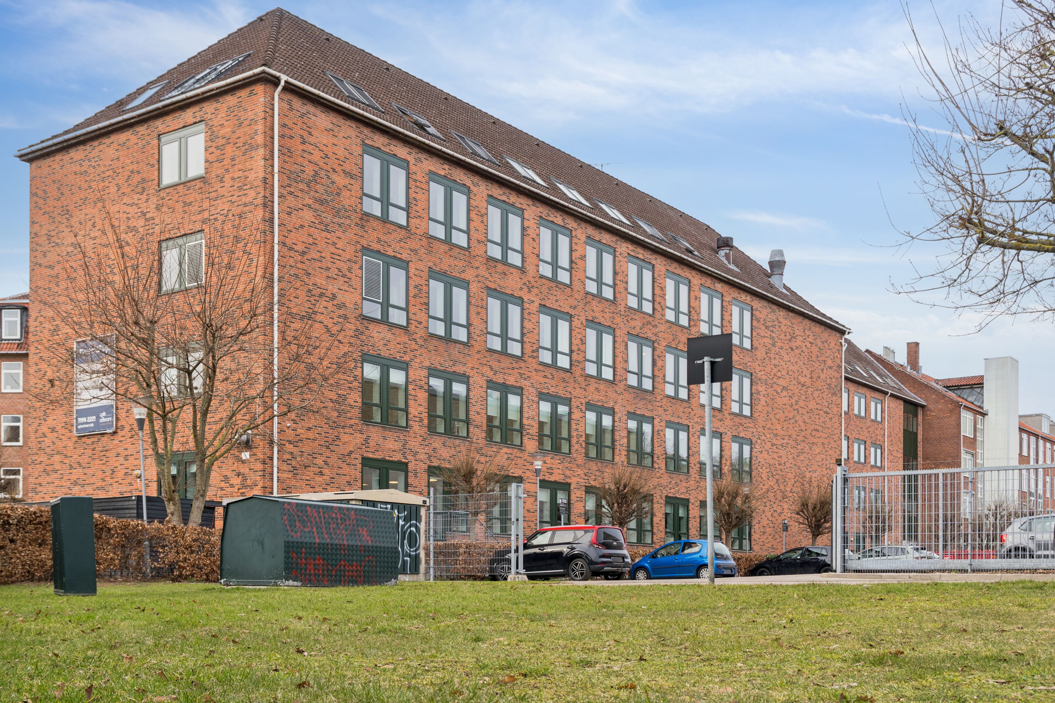 Lyst og indbydende kontorlejemål ved Nørrebro Station med udsigt til park, stående ved parkeringsplads med flere biler, røde teglstensbygning, vinduer og skur