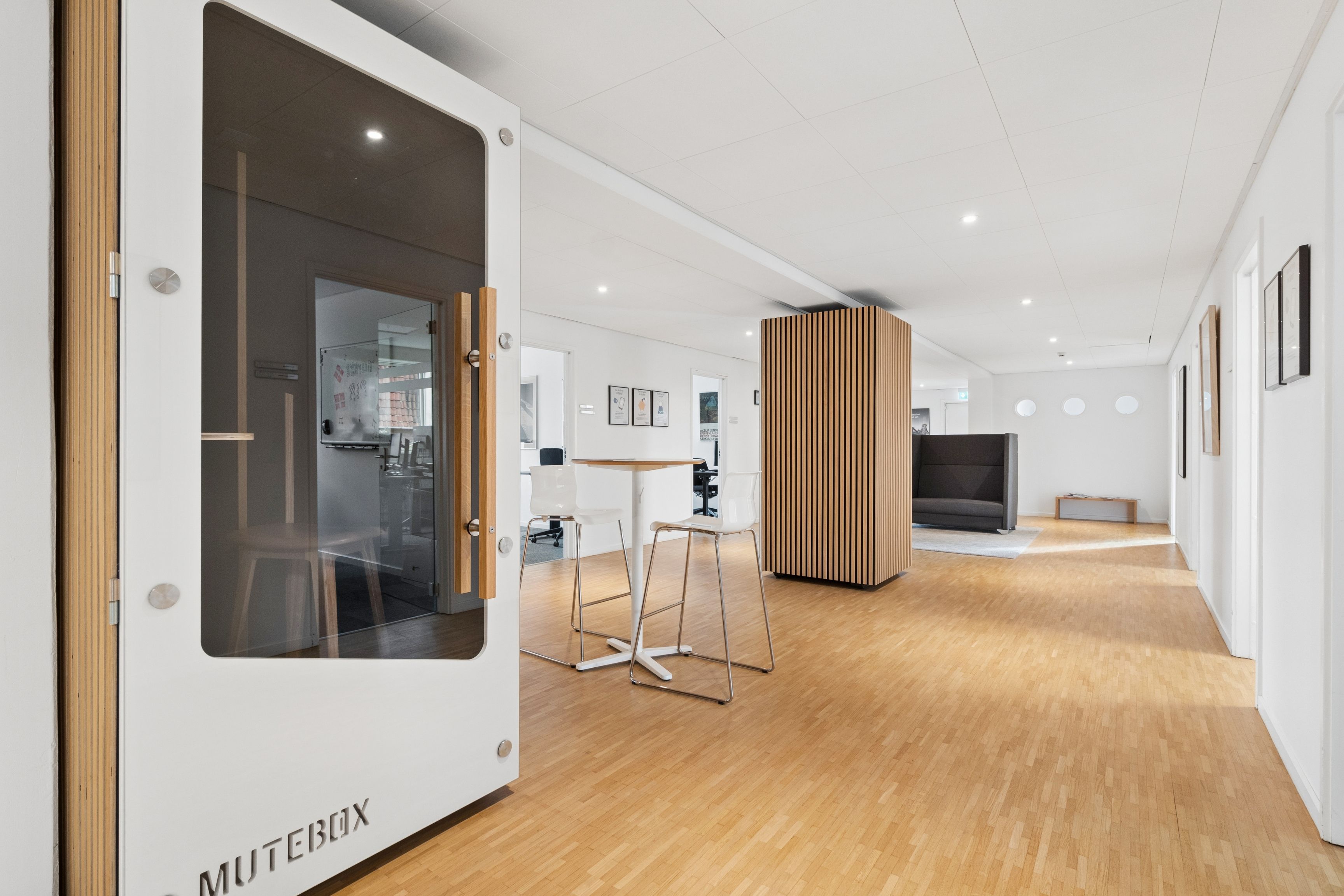 Kontorrum med moderne design, hvidt vægpanel og højt loft med indlejrede lyskatler. To hvide stole og et rundt bord står på trægulvet. Væggen er prydet med skrift og kunstværker.