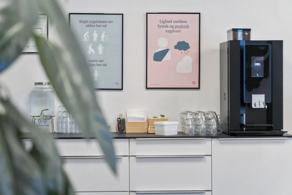Kontorlokaler med moderne interiør, to plakater med dansk tekst og et kaffeautomat udenfor. På væggen er der et kaffemaskine og en plante.