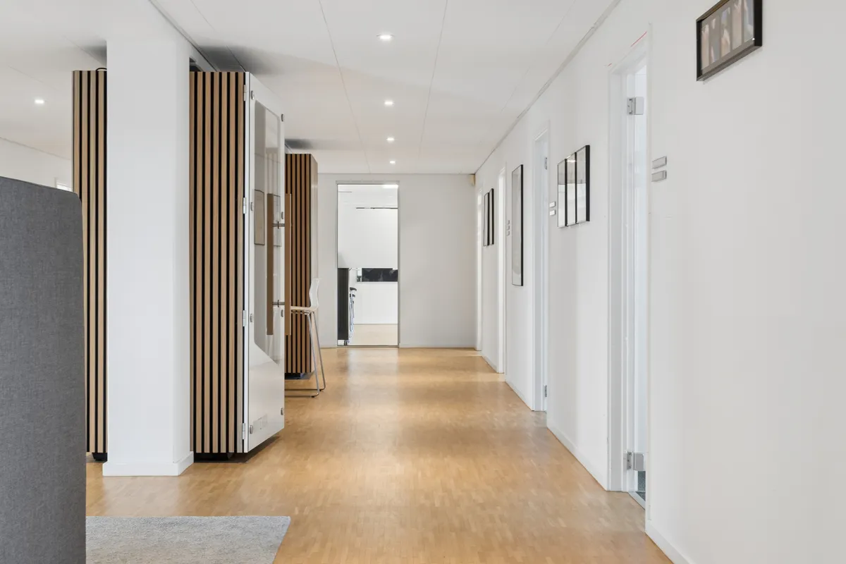 En lyse og minimalistisk korridor med lysegrå vægge, hvidt loft og lysebrune trægulve. Væggen er udsmykket med slanke, lyse møbler og tre små billeder